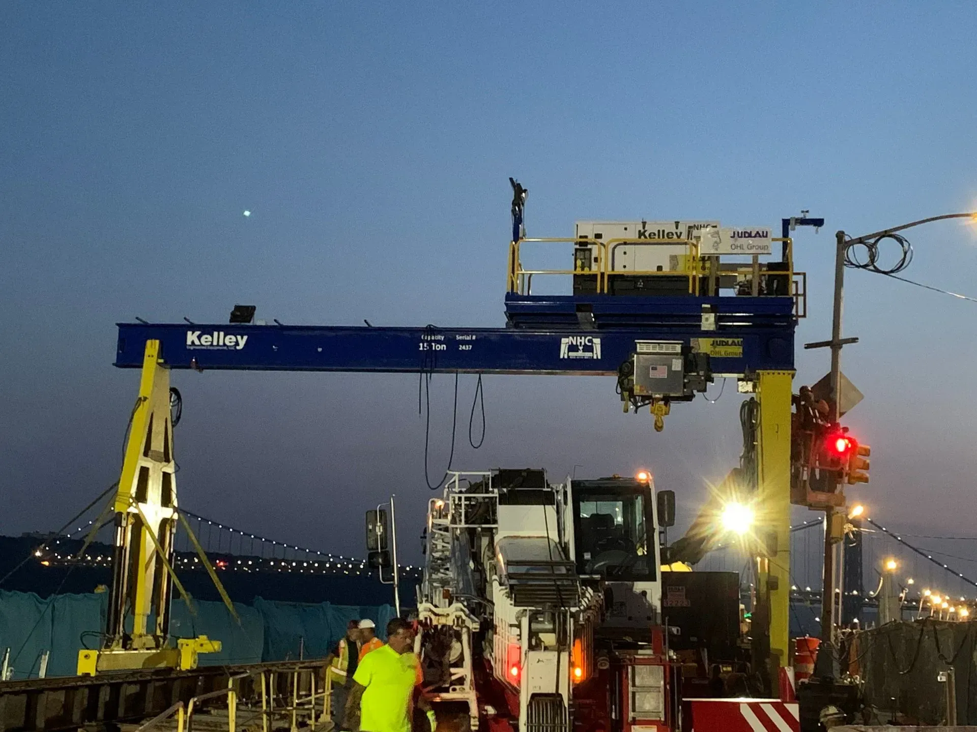 Crane lifting a white truck onto a flatbed at dusk, with workers and a red traffic light nearby.