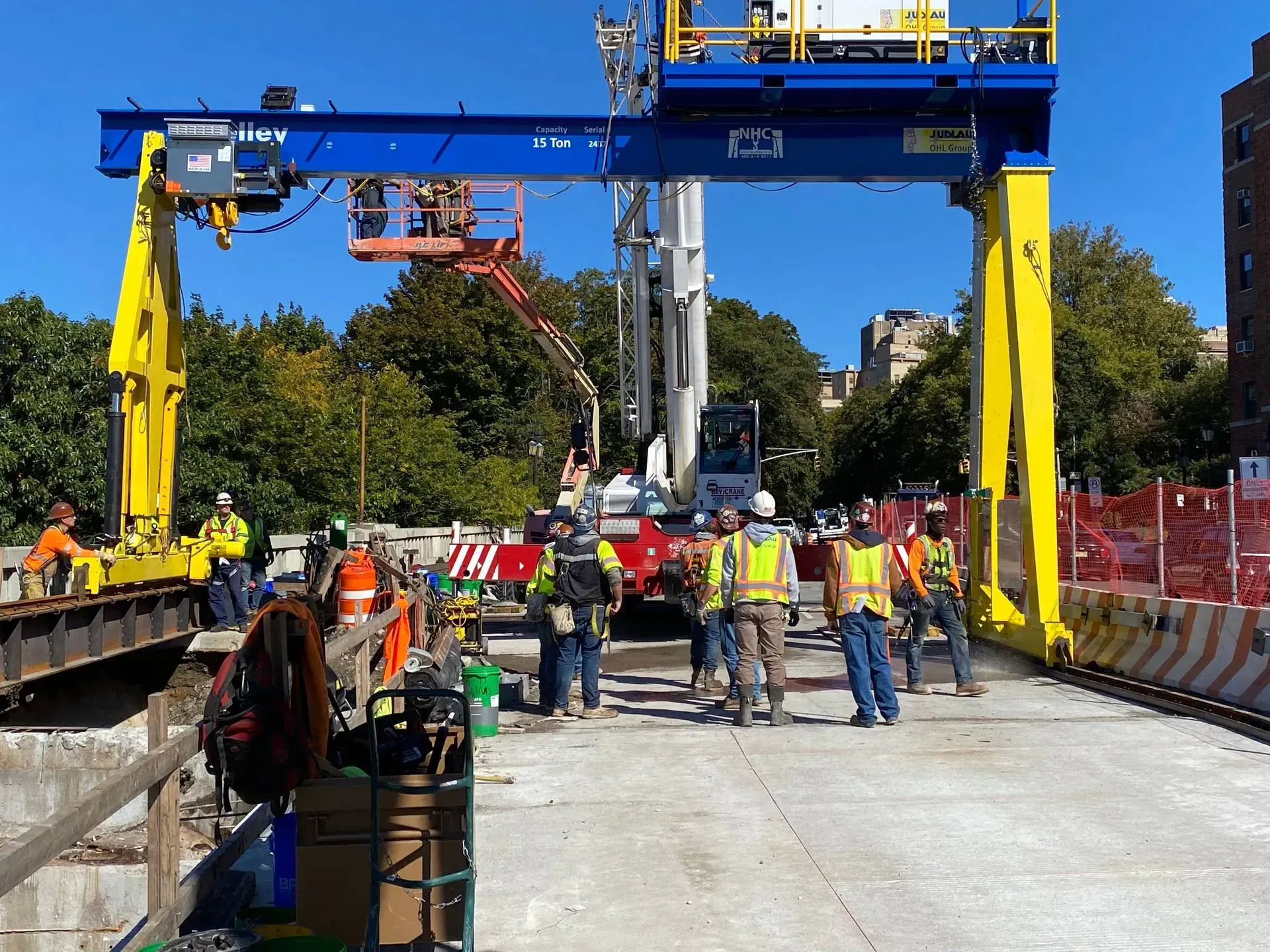 Workers on a construction site beneath a blue-and-yellow overhead crane, with barriers and equipment around a road trench.