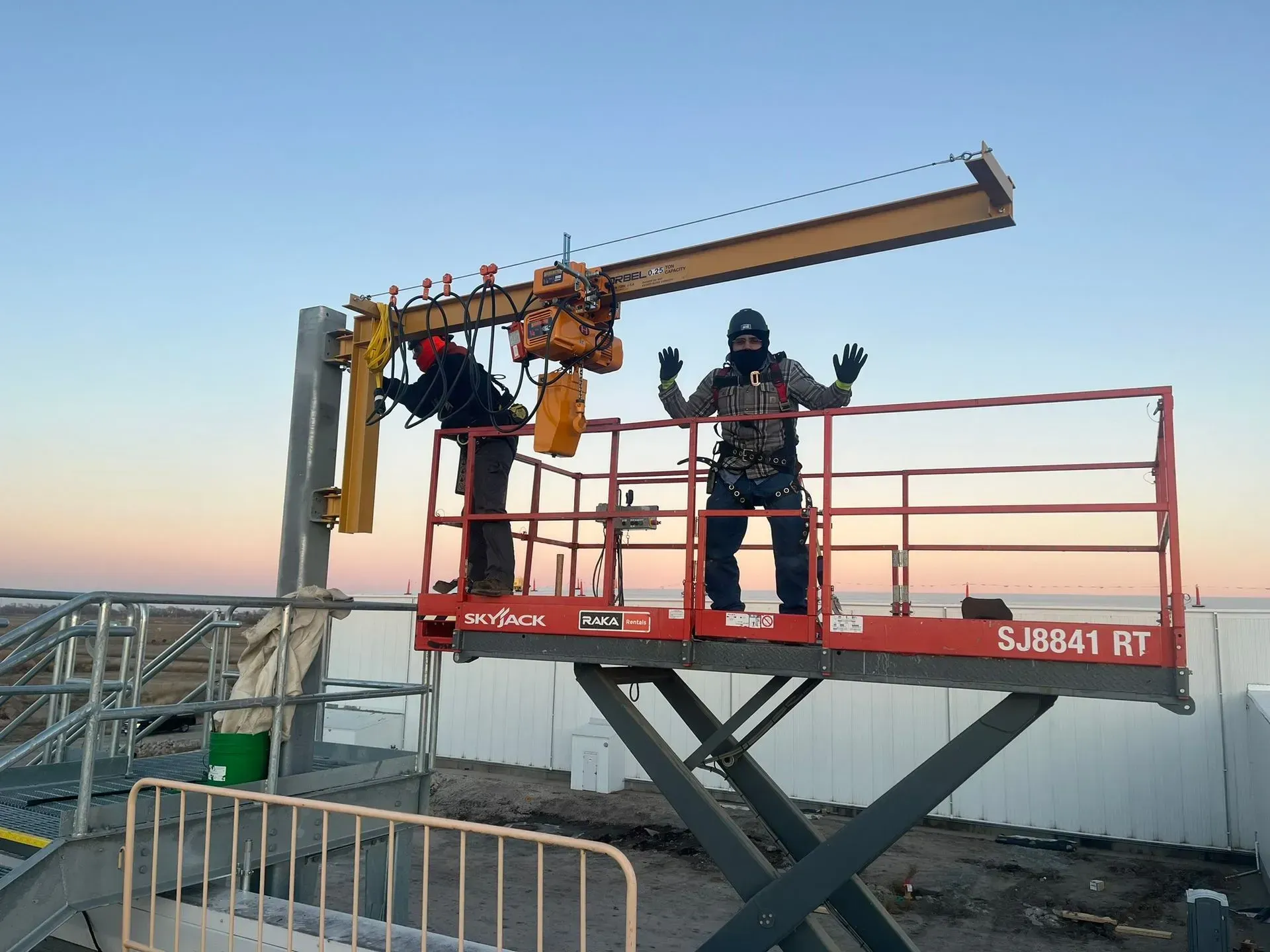 Two workers on a red lift platform inspecting a yellow industrial arm against a clear sky.