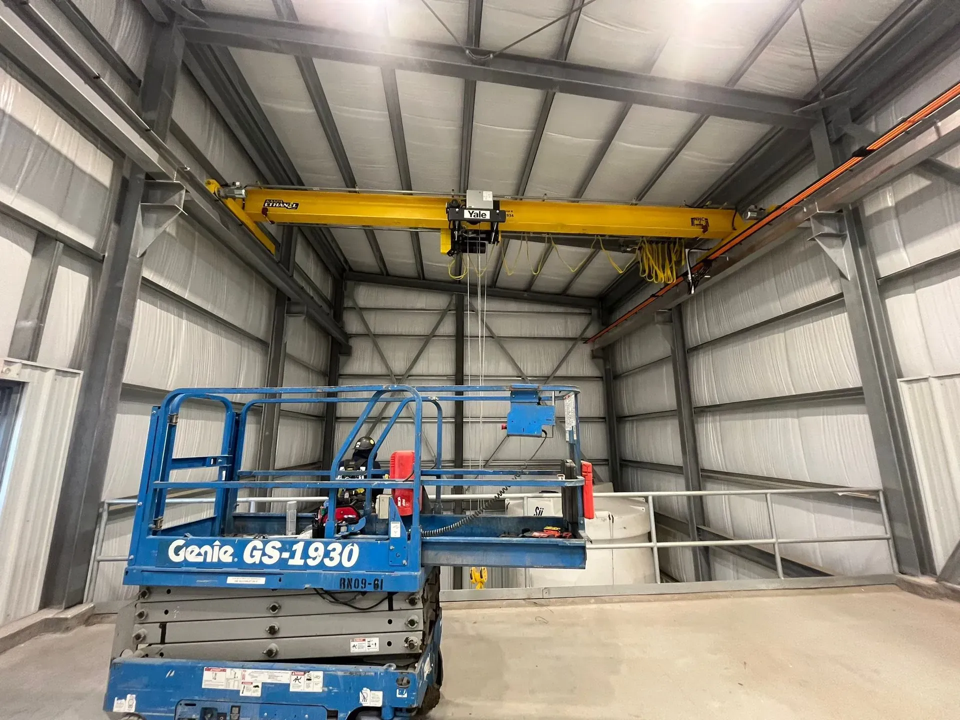 Blue Genie boom lift in a warehouse bay beneath an overhead yellow crane.