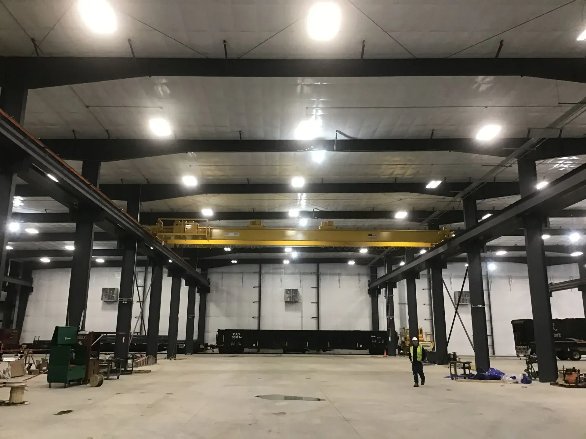 Large industrial warehouse interior with steel beams, bright overhead lights, and a few workers near equipment.