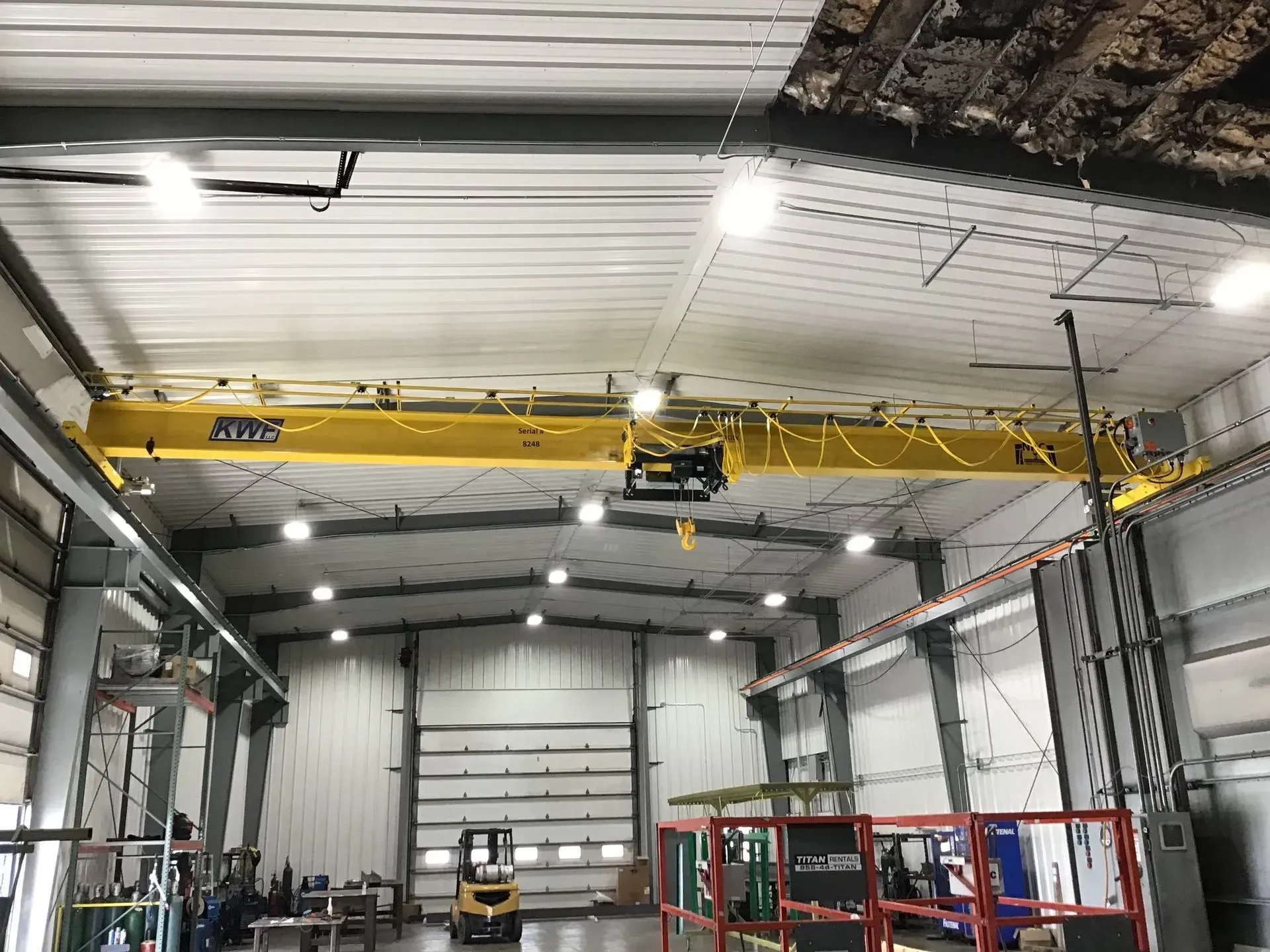 Industrial warehouse interior with yellow overhead crane, metal walls, and work vehicles below.