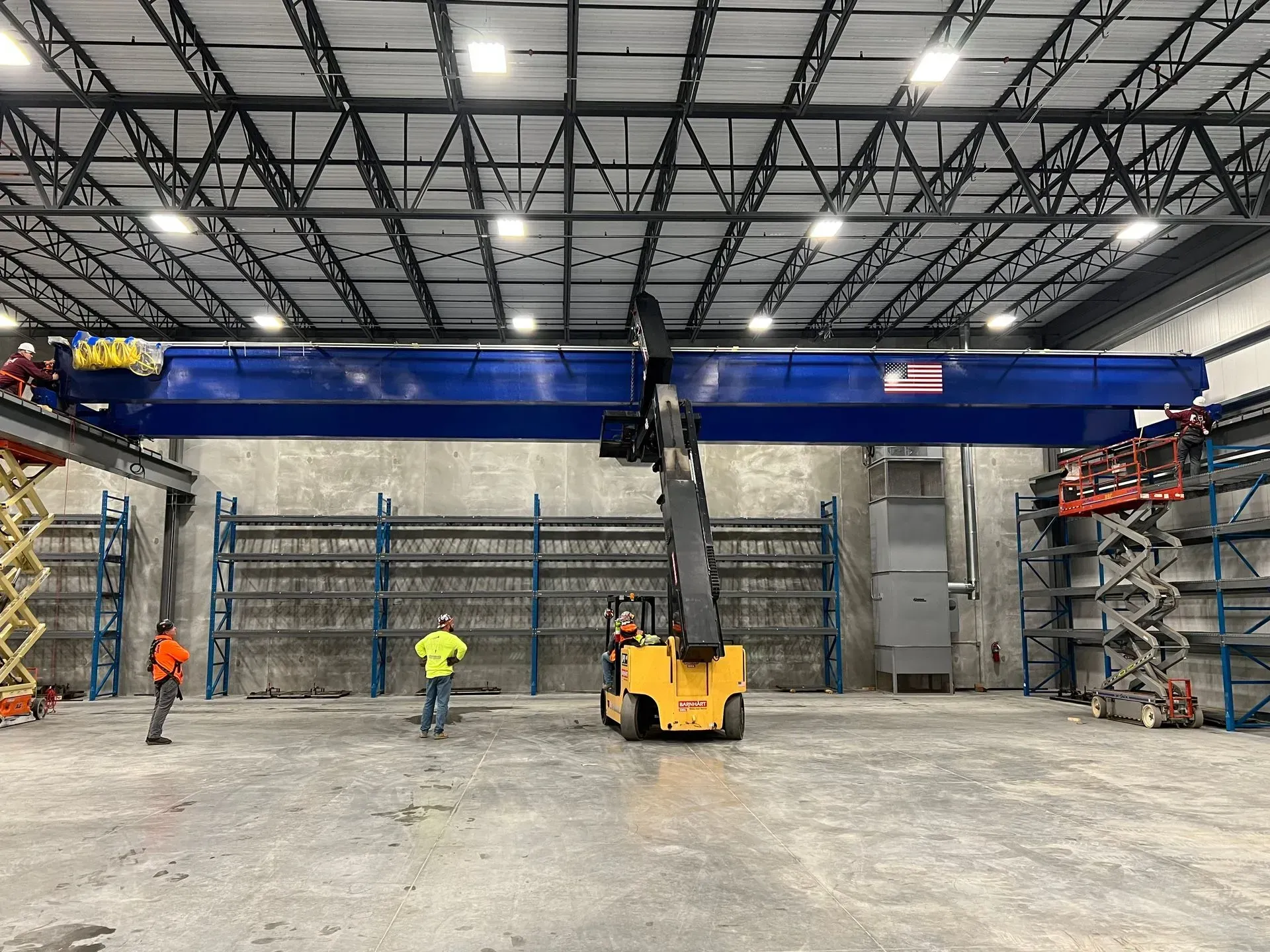Workers and a forklift installing a large blue overhead crane in a warehouse.