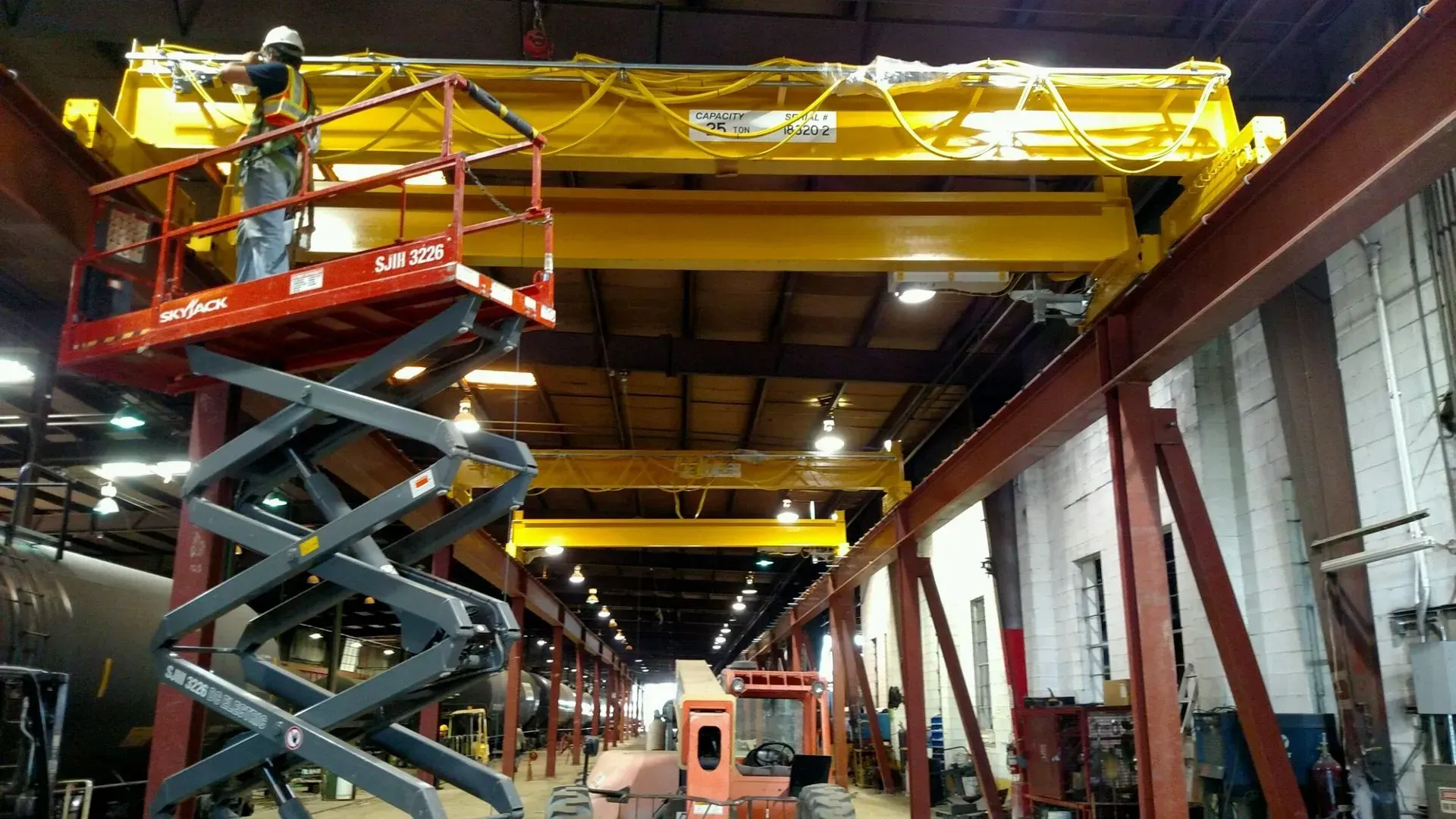 Worker on scissor lift inside industrial warehouse, servicing large yellow overhead crane.