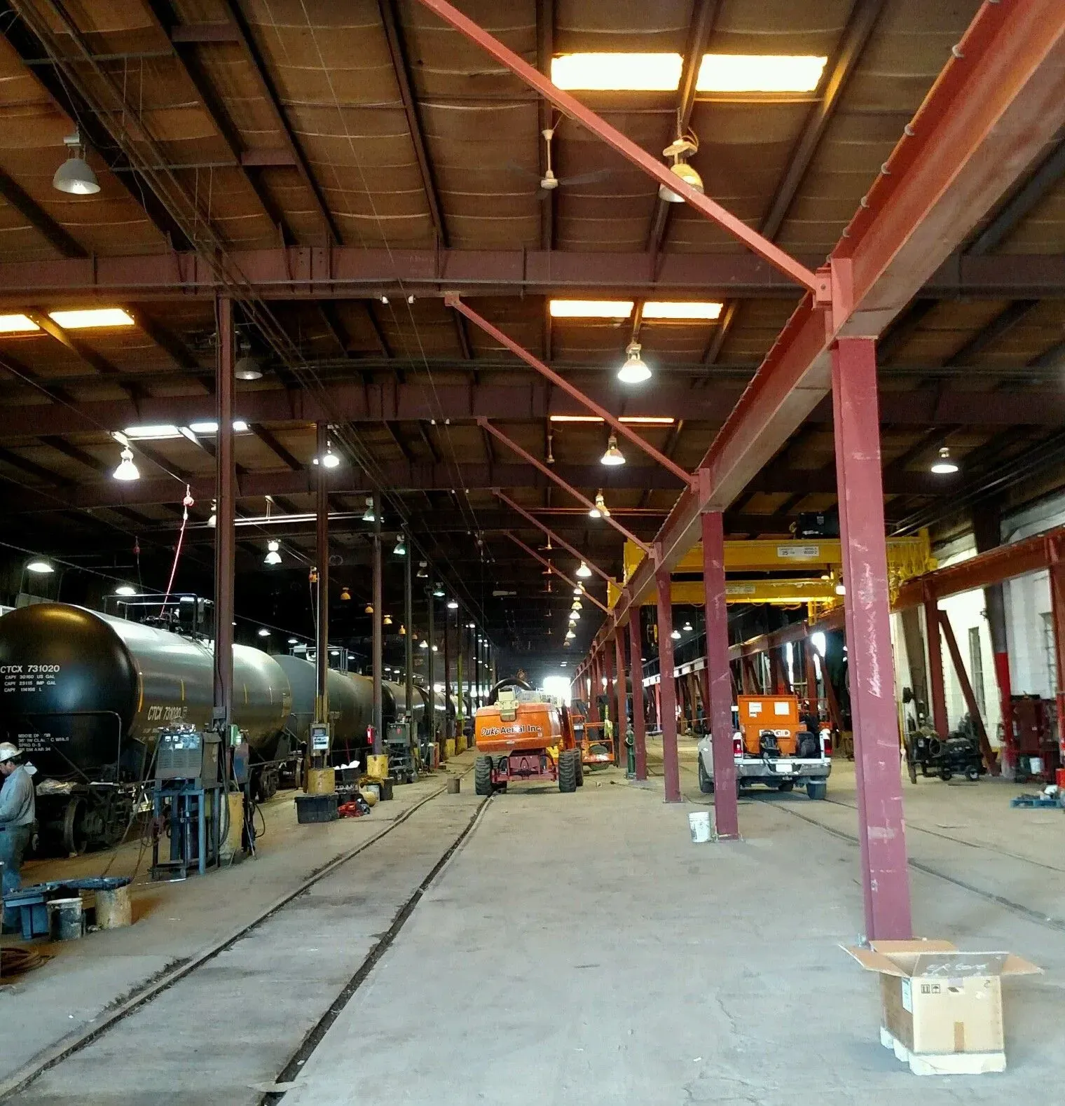 Interior of an industrial warehouse with trucks, machinery, and overhead lights on a concrete floor.