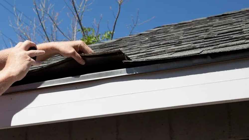 Hands lifting a shingle on a roof above a white gutter under a blue sky