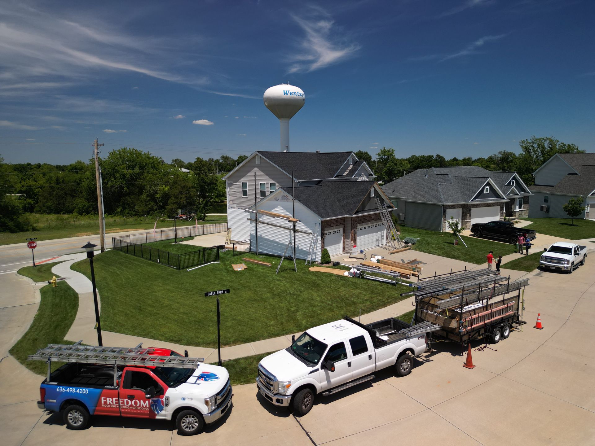 Workers and trucks at a house exterior renovation on a sunny suburban street