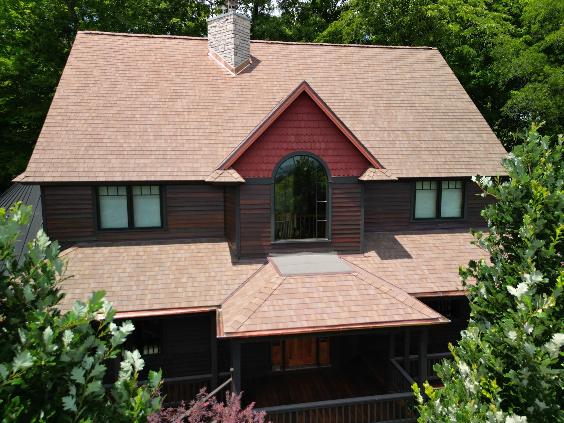 Two-story house with brown siding, tan roof, and red gable among dense green trees