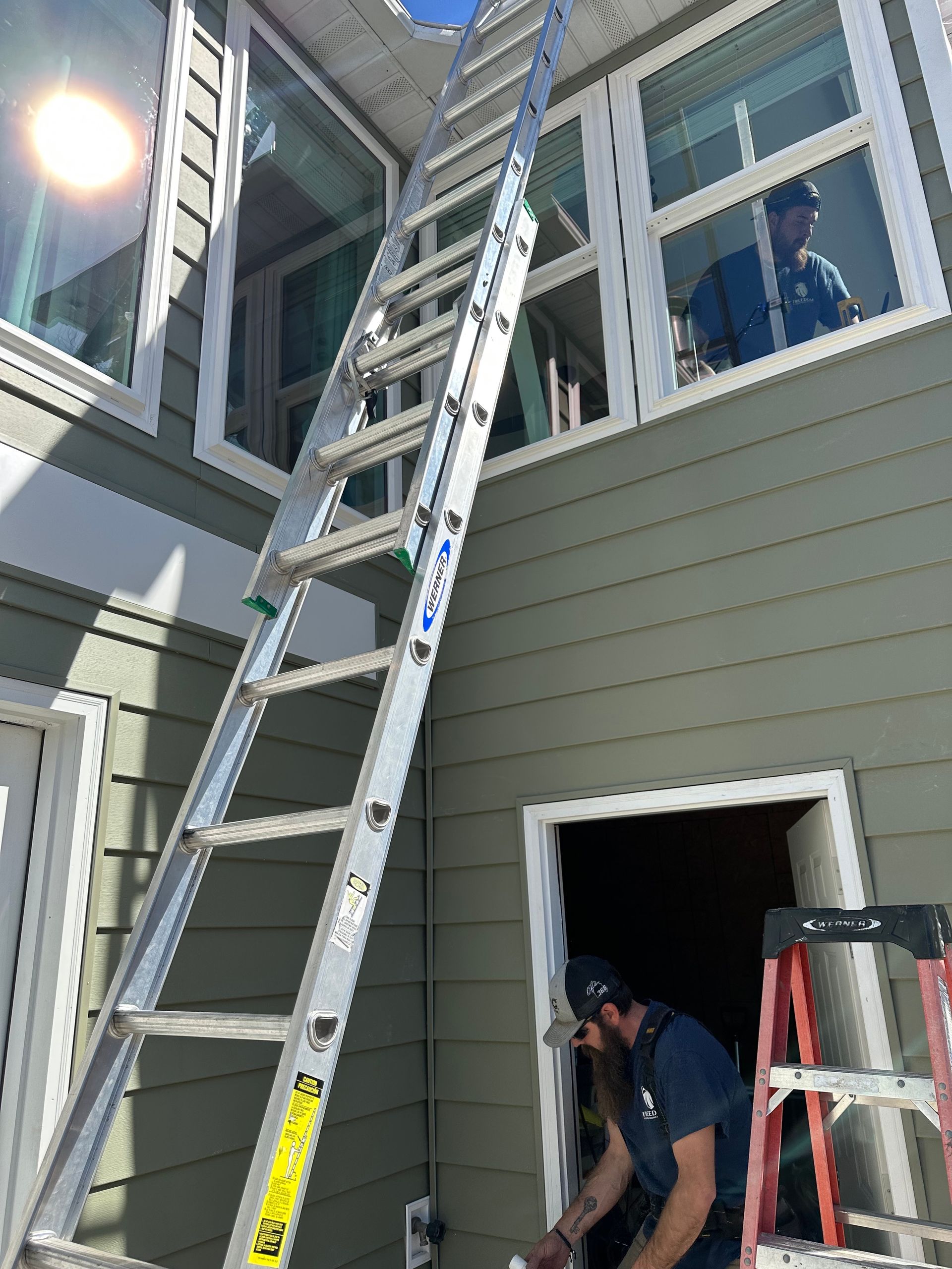 Two workers on ladders outside a house, working near an upper window and an open doorway.