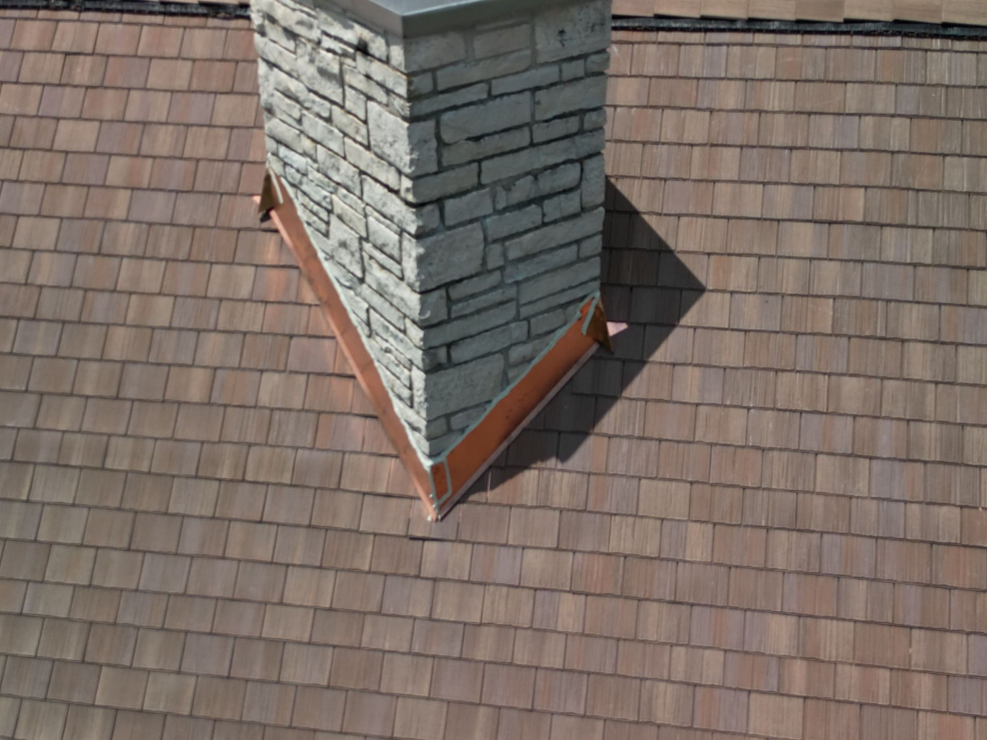 Rooftop chimney with gray stone siding and copper flashing on brown shingle roof