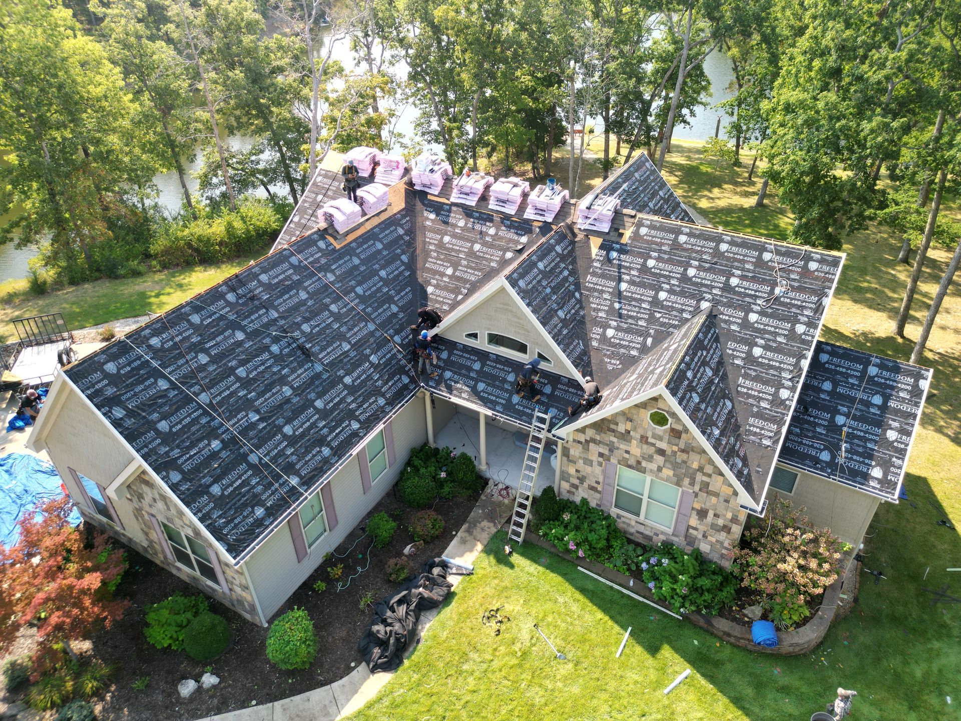 Aerial view of a house with a dark roof, stone exterior, and surrounding green trees and lawn.