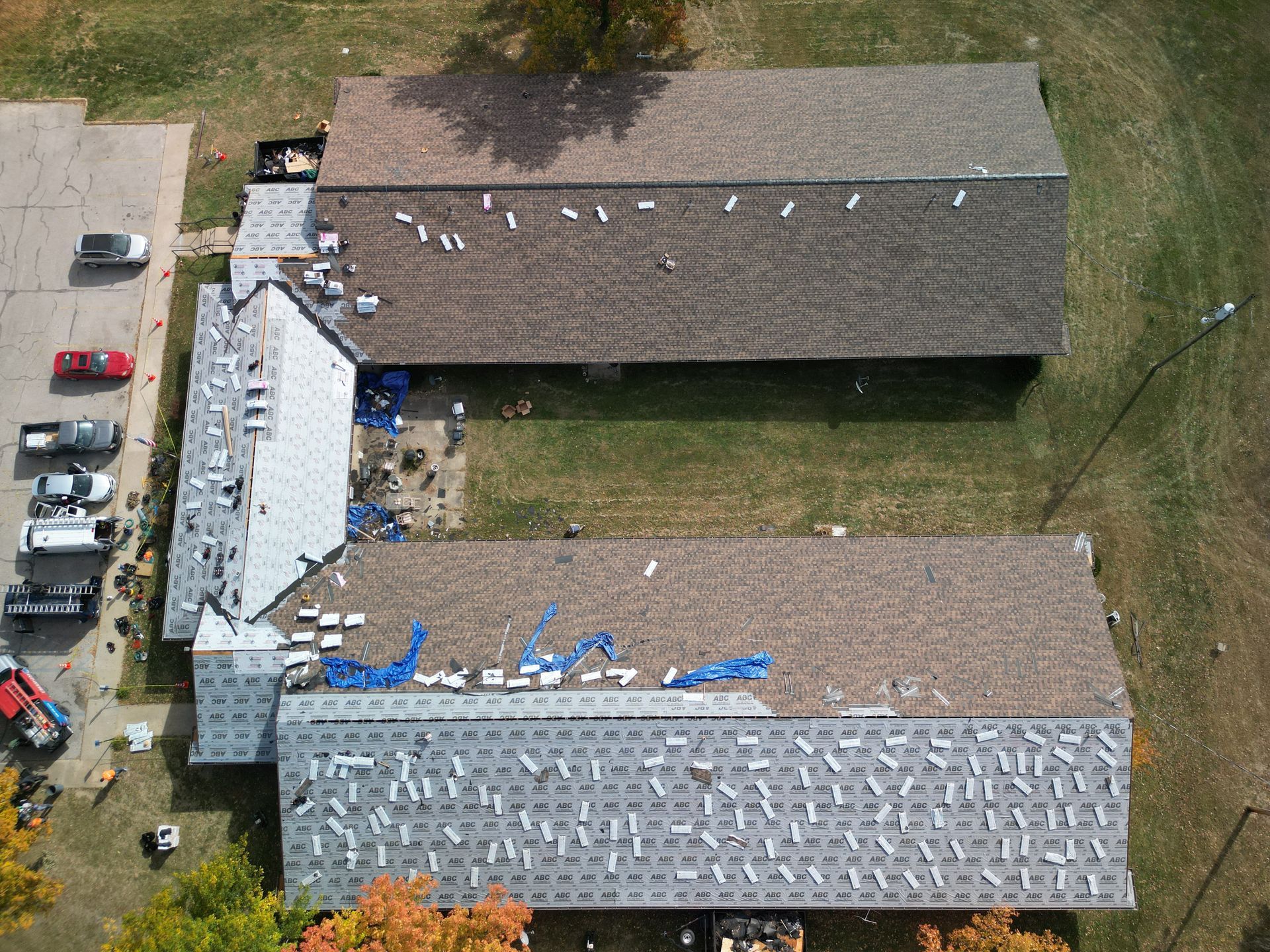 Aerial view of two long buildings with damaged roofs, debris, and construction equipment beside a grassy lot.