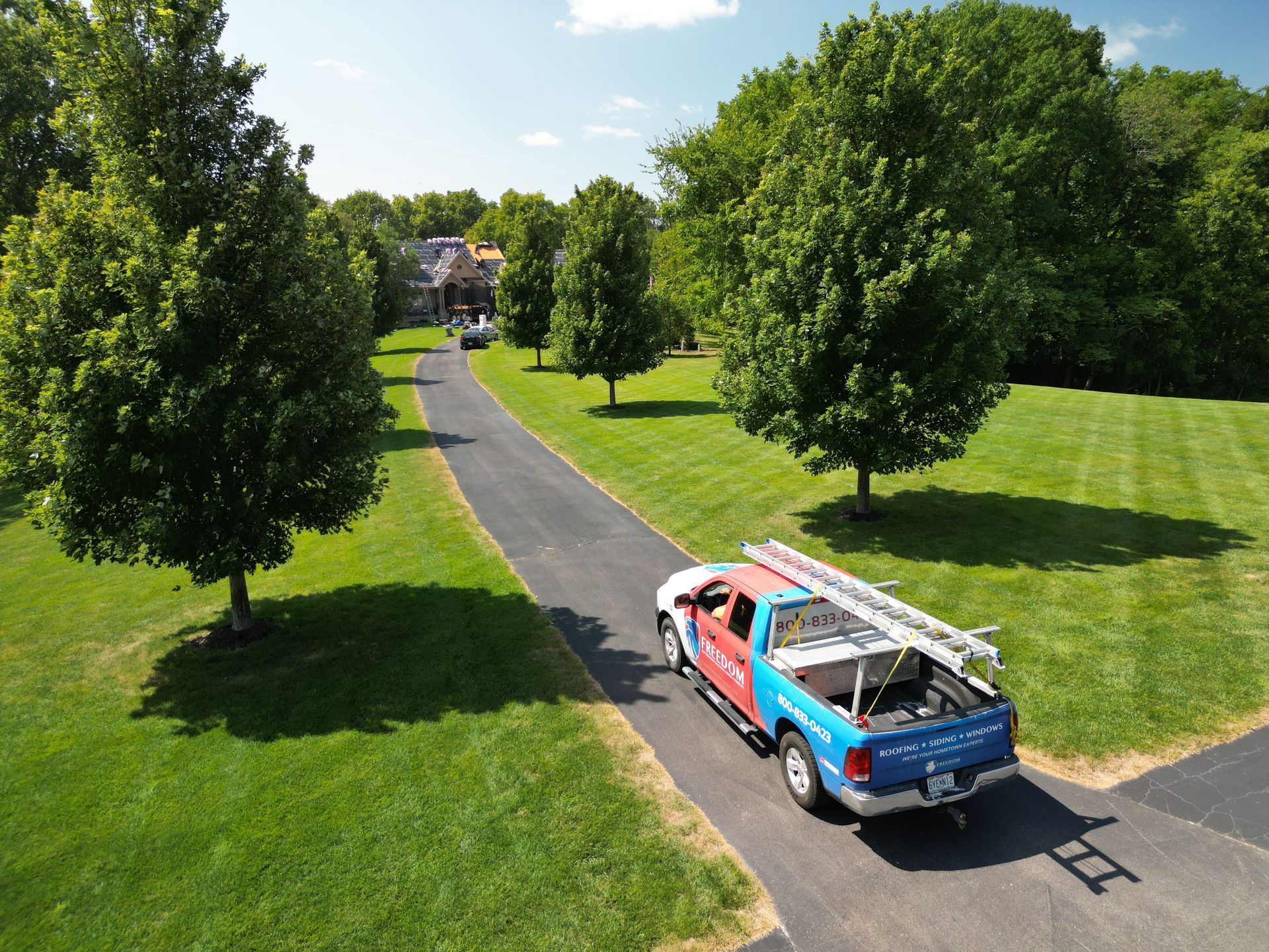 Blue utility truck on a park road beside green lawns and trees, near a distant building