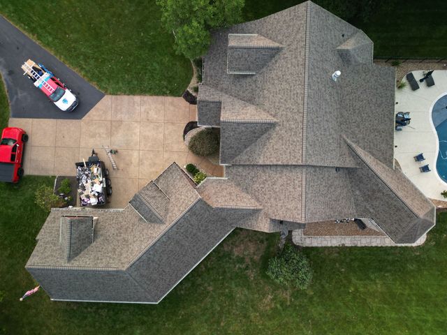 Aerial view of a large gray-roofed house with driveway, patio, and parked vehicles surrounded by grass.