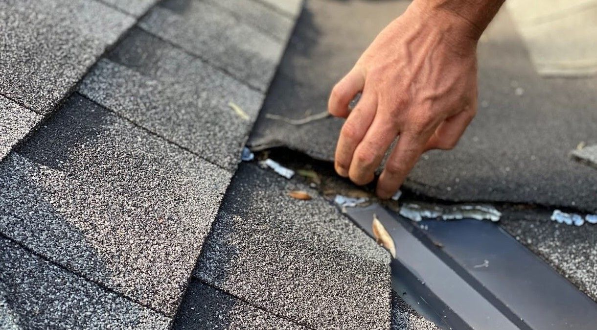 Hand inspecting a damaged asphalt shingle roof with exposed underlayment and flashing