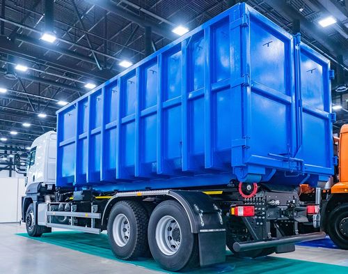 Blue dump truck in a bright warehouse, shown from the rear side.