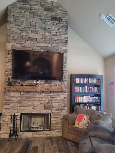 Living room with stone fireplace, wall-mounted TV, bookshelf, and beige chair with red dollhouse toy.