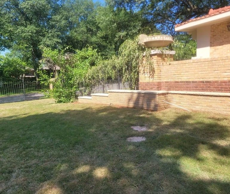 Backyard lawn beside a brick house, with trees and a low retaining wall in bright sunlight