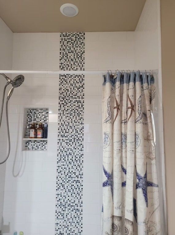 Bathroom shower with white tile, mosaic accent strip, built-in shelf, and patterned curtain.