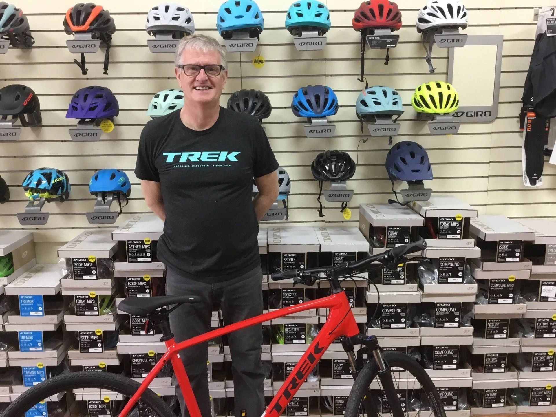 Man in Trek shirt standing next to a red bike in a bike shop. Helmets displayed on wall behind him.
