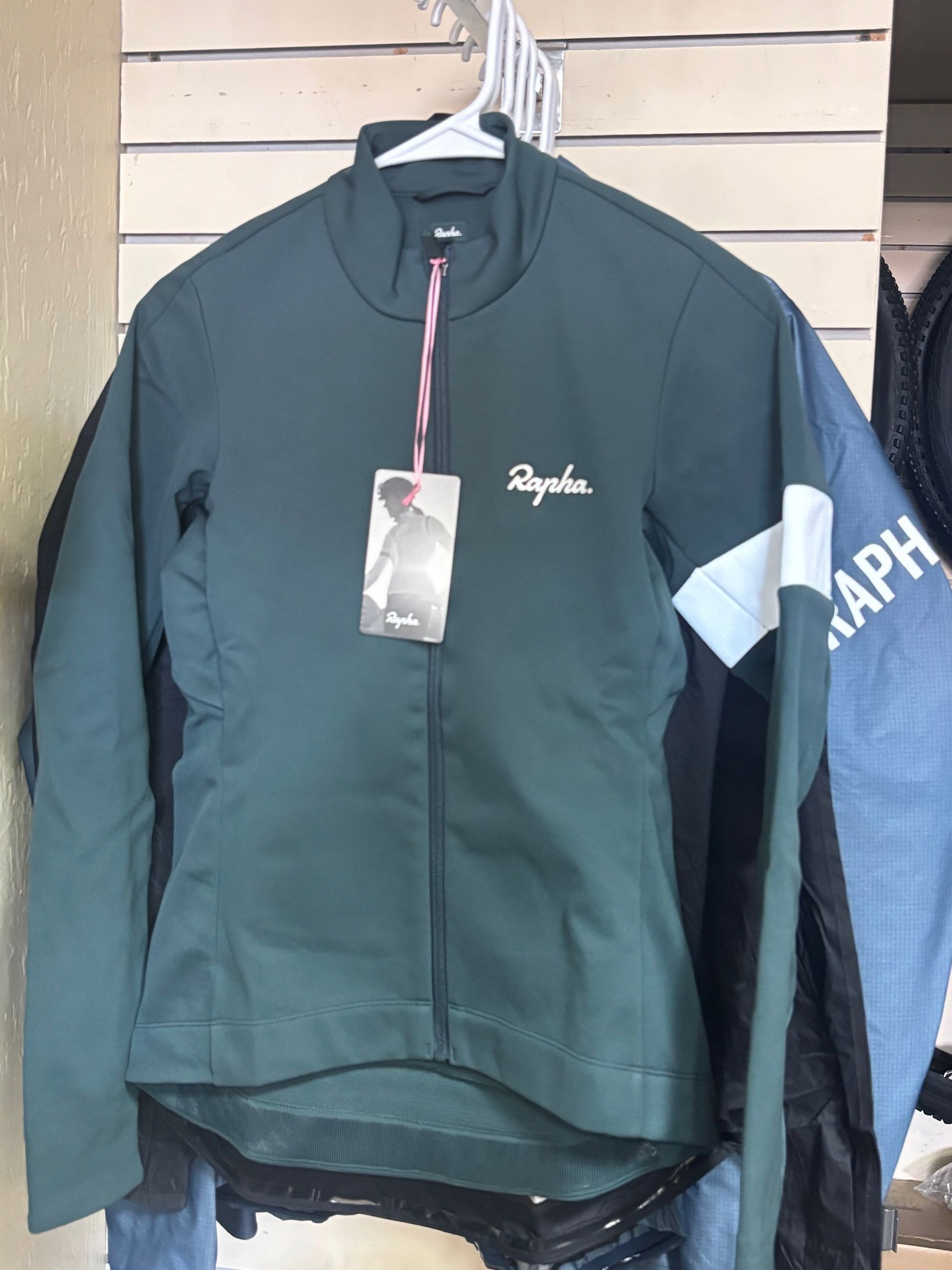 Green Rapha cycling jacket with white logo and tag, hanging in a store.