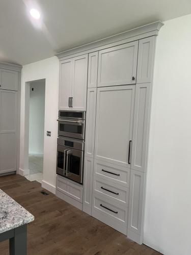 A gray kitchen cabinet with built-in oven, microwave, and refrigerator with black handles and white walls.