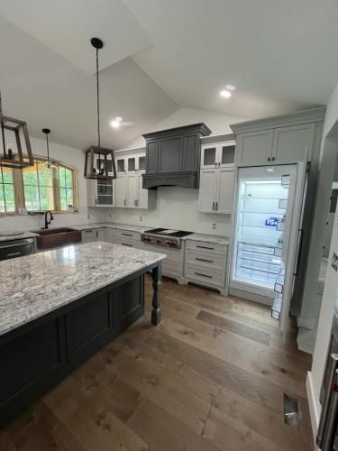 A modern kitchen with light gray cabinets, dark island, and granite countertops. Light oak flooring and open refrigerator visible.