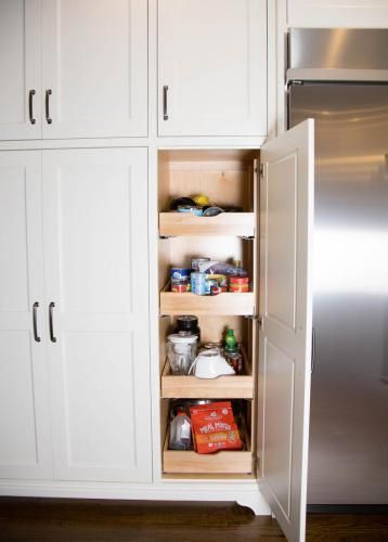 A white kitchen pantry cabinet with pull-out shelves, stocked with food items, next to a stainless steel refrigerator.
