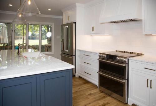 Modern kitchen with a blue island, white cabinets, stainless steel appliances, and a large window overlooking a green lawn.