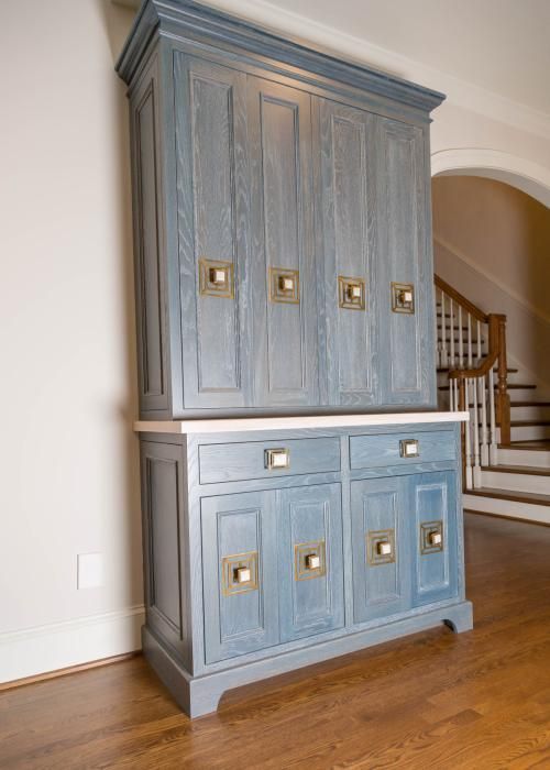 Blue wooden cabinet with brass hardware, set against a beige wall and hardwood floor, with a staircase visible in the background.