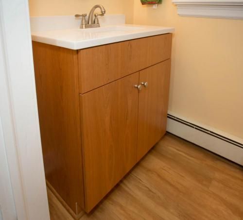 A light wood bathroom vanity with a white countertop, sink, and silver faucet. It sits against a yellow wall with a brown floor.