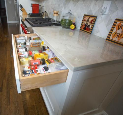 A kitchen island with a spice drawer open, showing various spice containers. White countertops, cabinets, and backsplash with Halloween decorations in view.