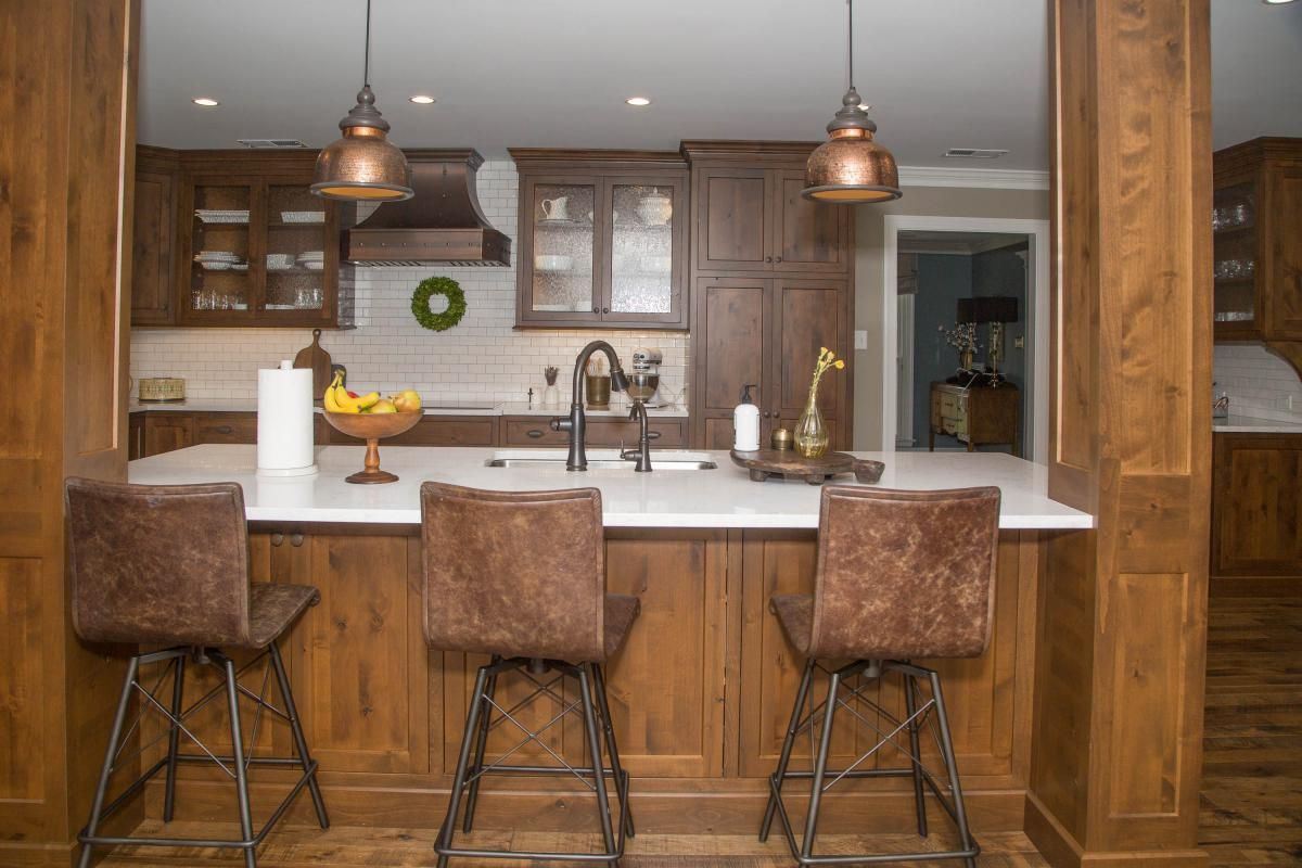 A modern kitchen with dark wood cabinetry, a white countertop island with three brown leather bar stools, and copper pendant lights.