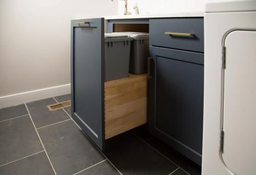 Pull-out cabinet in a laundry room with two trash bins. Dark blue cabinets, gray bins, and dark gray tiled floor.