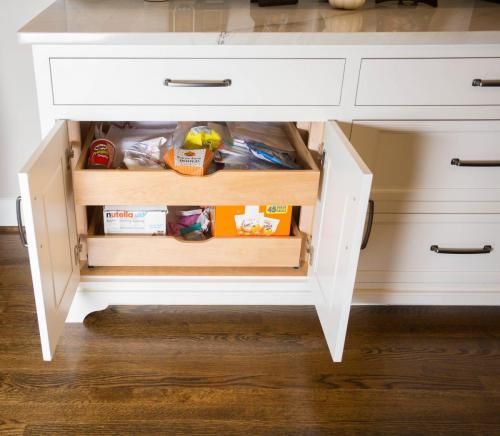 White kitchen cabinet with open doors revealing pull-out drawers holding packaged food items.  Dark hardware and a wood floor.