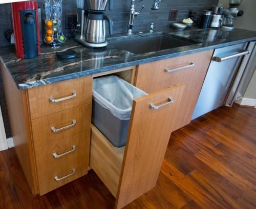 A wooden kitchen cabinet with pull-out trash bin and drawers; countertop sink and dishwasher in the background.