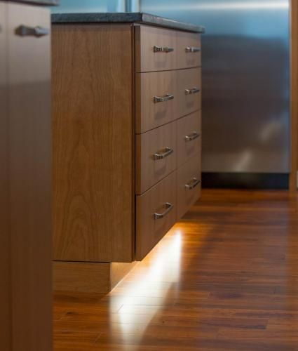 Wooden kitchen cabinet with drawers. Warm lighting illuminates the floor and cabinet base.