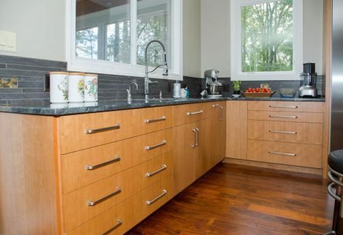 A modern kitchen with light wood cabinets, dark countertops, and large windows. A stainless steel faucet is above the sink.