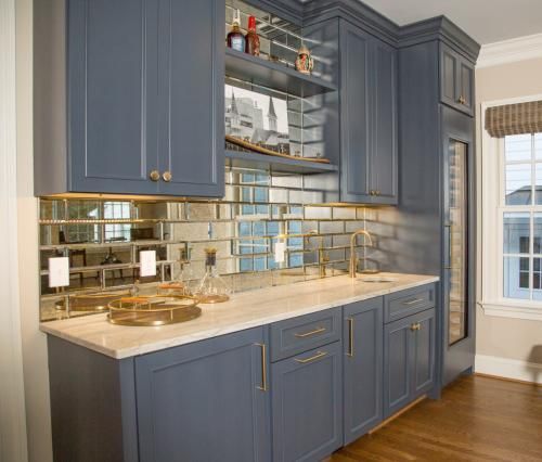 A blue built-in home bar with a mirrored backsplash, countertop, and cabinets. A wine fridge and window are nearby.