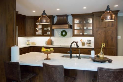 A modern kitchen with dark wood cabinets, white countertops, and copper pendant lights. There are kitchen bar stools and a centerpiece on the counter.