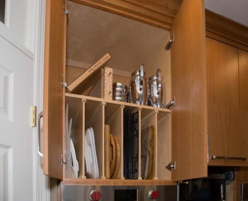 Open kitchen cabinet with dividers organizing baking sheets and utensils. Light wood with a stainless steel oven below.