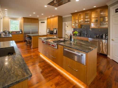 Spacious kitchen with wood cabinets, granite countertops, and stainless steel appliances. Island features a cooktop and dishwasher, lit by under-cabinet lights.