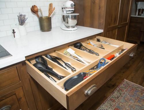 Kitchen drawer filled with utensils and small tools, with wooden dividers for organization. Brown cabinetry and white countertop.