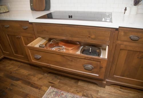Wooden kitchen cabinets with a drawer open, revealing cookware. Light wood floor, white countertop, and a stovetop are visible.