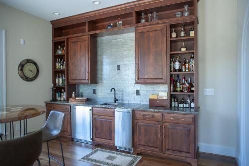 A home bar area with dark wood cabinets, stainless steel appliances, and a decorative tiled backsplash. There is a sink, shelving with liquor bottles, and a small rug.