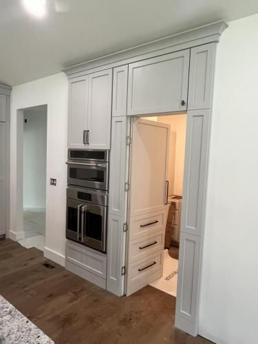 A gray cabinet unit in a kitchen concealing a hidden door. It houses an oven and microwave. The door reveals a small room.