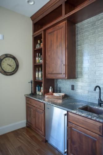 Built-in bar area with dark wood cabinets, a stainless steel mini-fridge, and a brick-patterned backsplash. Includes a sink and a wall clock.