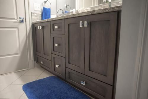 A dark wood bathroom vanity with two cabinet doors, drawers, and silver hardware. A blue bathmat is on the tiled floor.