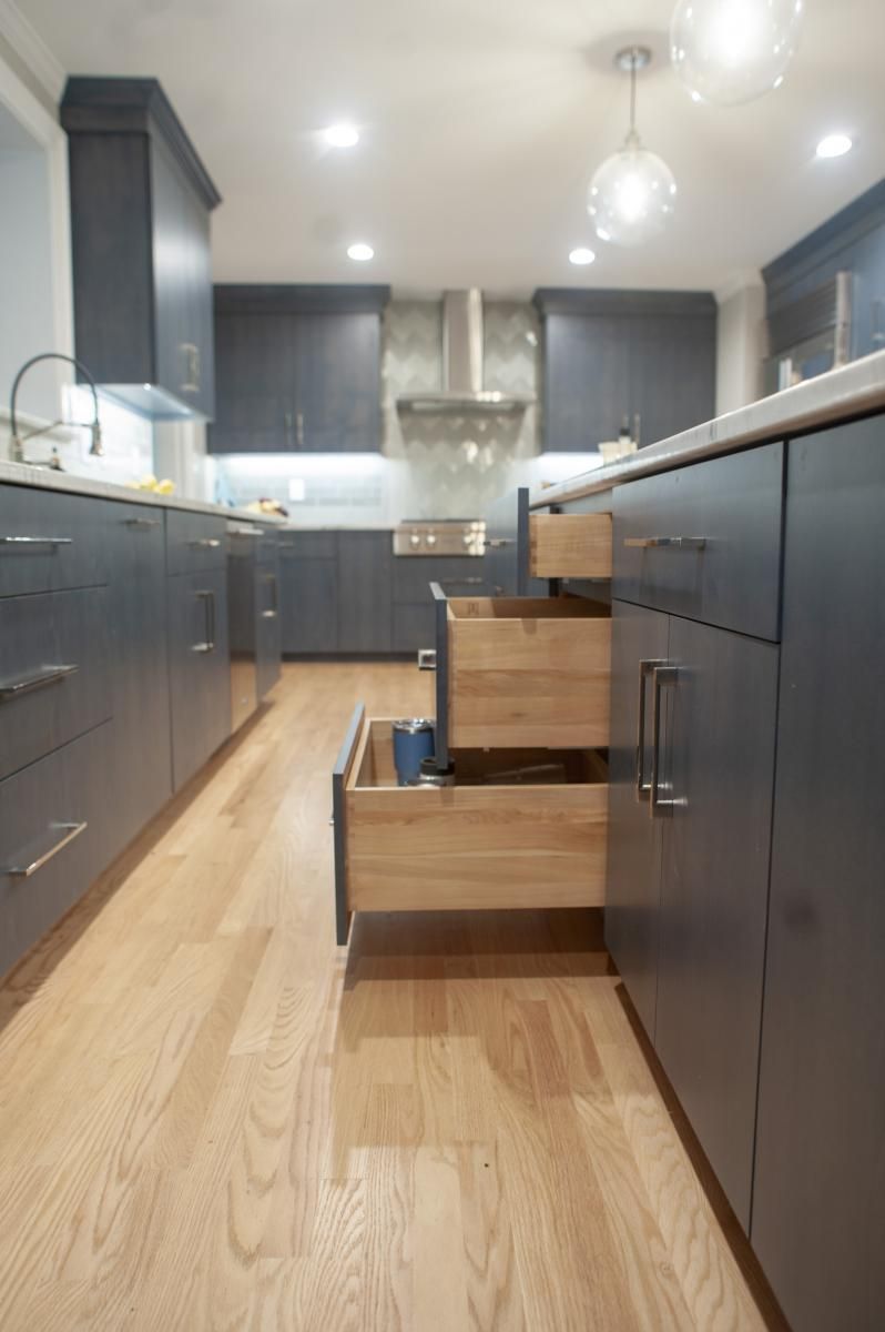A modern kitchen with dark blue cabinets and light wood floors. Two open wooden drawers reveal kitchenware.