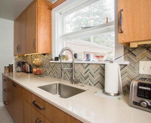 A kitchen sink area with light wood cabinets, white countertops, and a patterned backsplash. A window and a toaster are also visible.